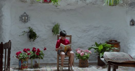 Dor e Glória (Dolor y Gloria), de Pedro Almodóvar. Cena com o menino Asier Flores sentado na cadeira, paredes brancas e flores atrás, lendo um livro.