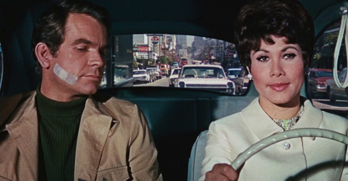 Personagens principais do filme "Se Meu Fusca Falasse" - a mulher está no volante e o homem está olhando para ela com um curativo na bochecha