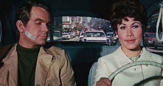 Personagens principais do filme "Se Meu Fusca Falasse" - a mulher está no volante e o homem está olhando para ela com um curativo na bochecha